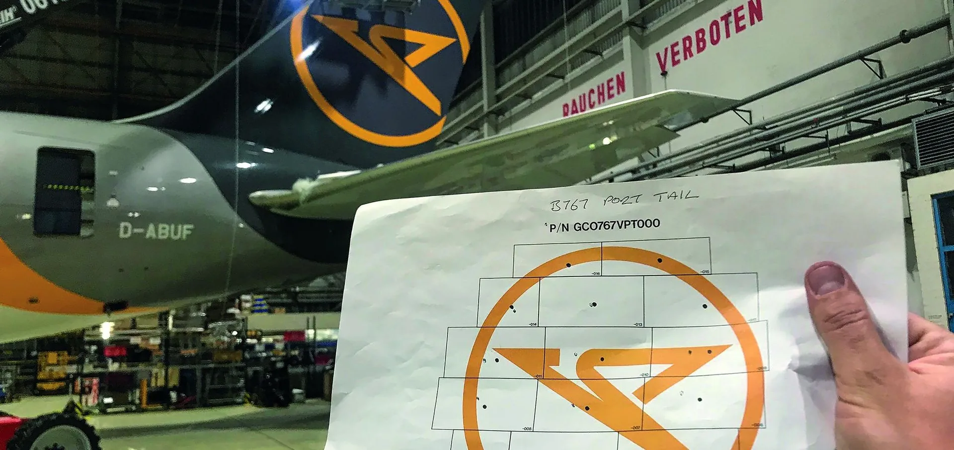 Printed tail template marked B767 port tail with its part number, held up in front of the aircraft in the hangar